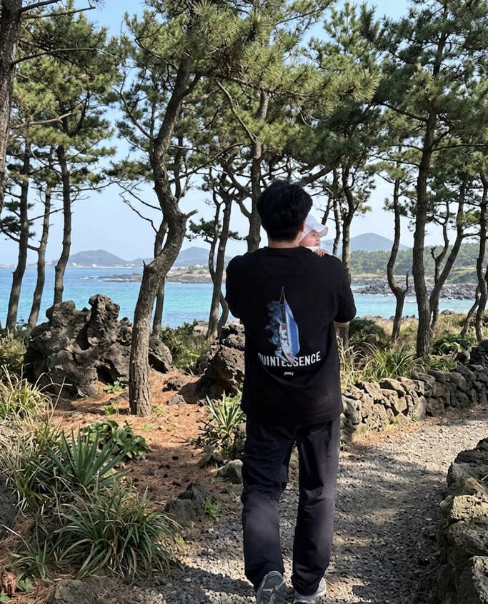 푸른 제주 바다와 현무암이 어우러진 해안 산책로를 배경으로, 아빠가 어린 딸을 소중하게 품에 안고 숲길을 걷고 있는 뒷모습. 평화로운 가족 여행의 한 장면